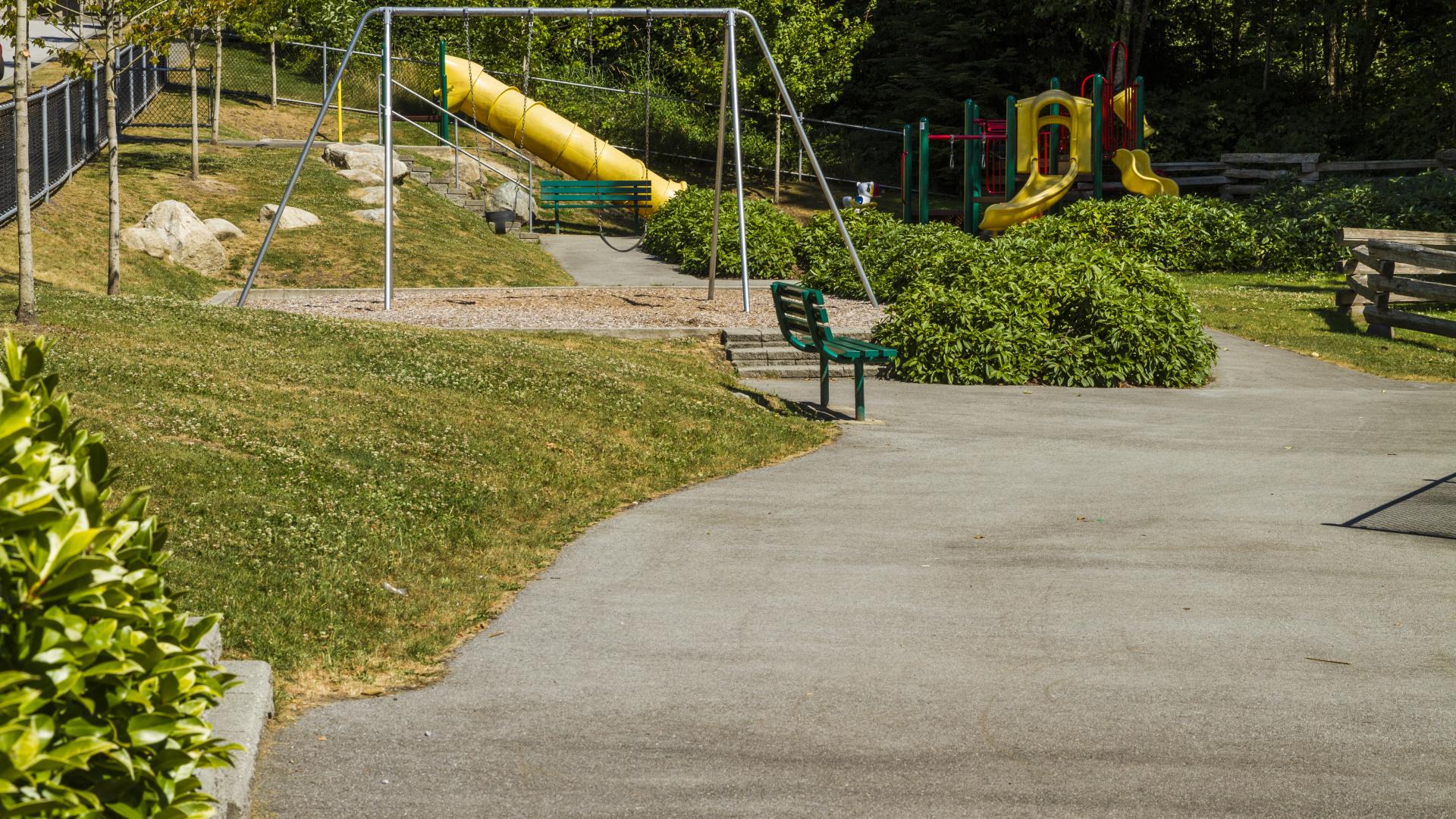 A wide paved pathway leading toward a park bench, a swing set, and, beyond, a playground.
